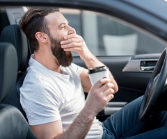 A man yawning in his car while holding coffee.